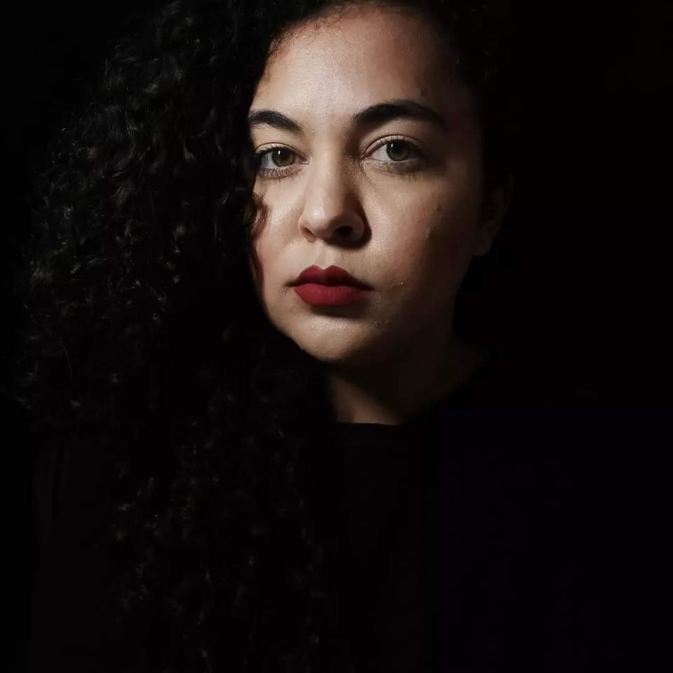Menina de olhos esverdeados com cabelo castanho escuro cacheado, esta com uma blusa preta e batom vermelho.
